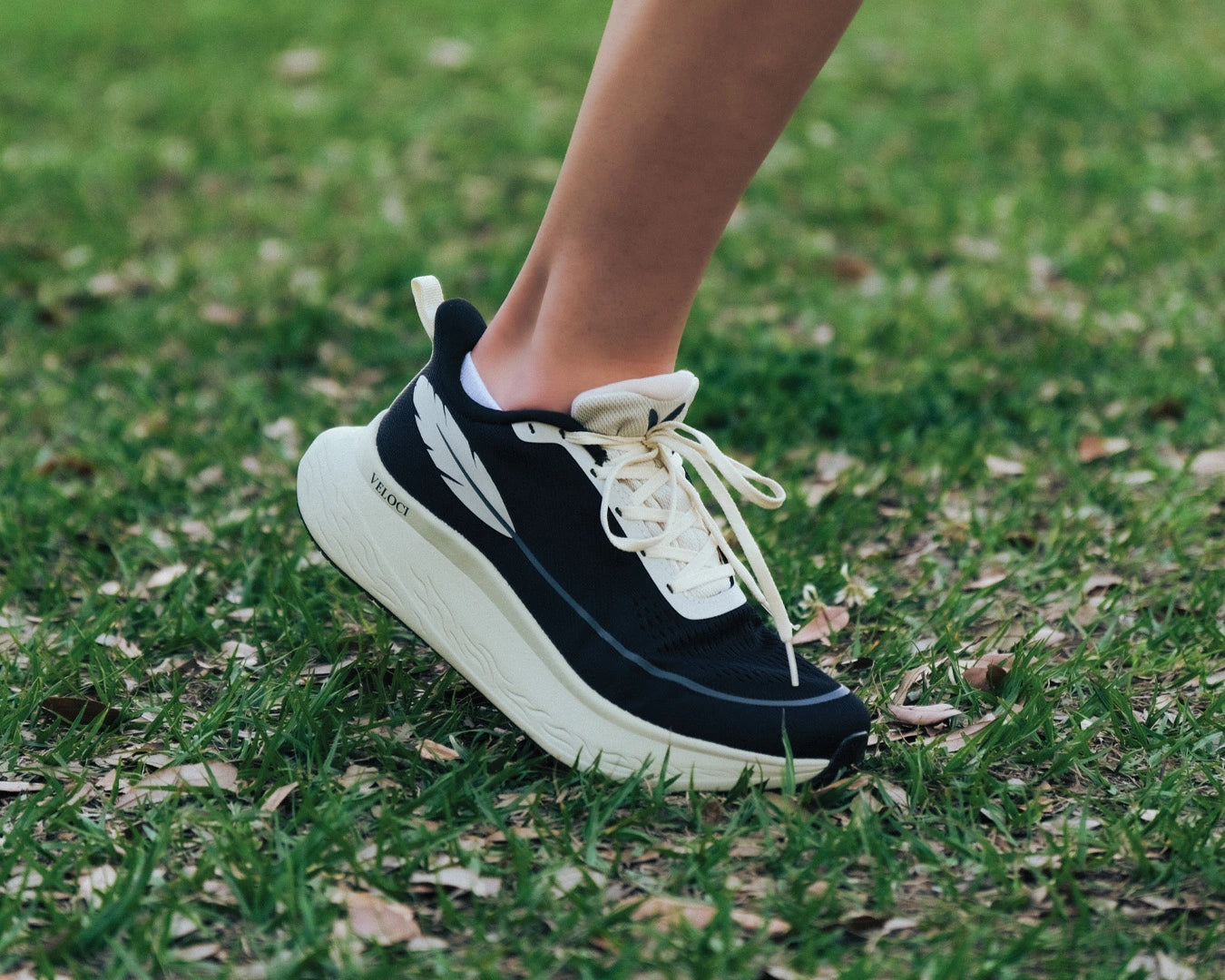 Veloci Ascent running shoe in black with cream midsole, outdoor grassy side profile showing the supportive foam, wide toe box, and breathable mesh upper